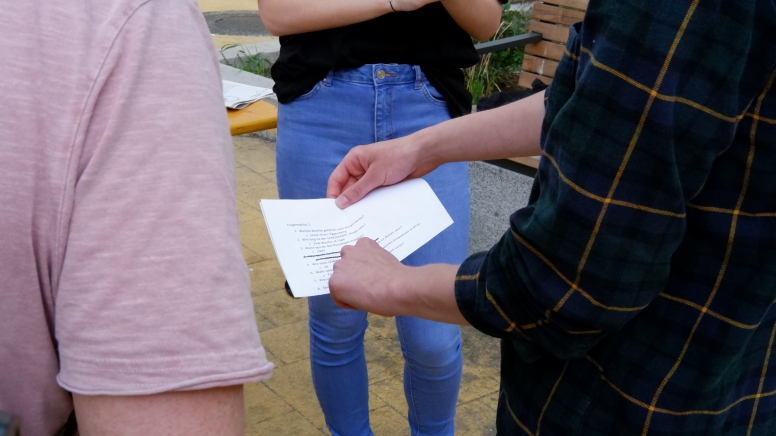 Eine Hand Zeigt auf einen zettel mit Quizfragen, daneben stehen zwei Menschen