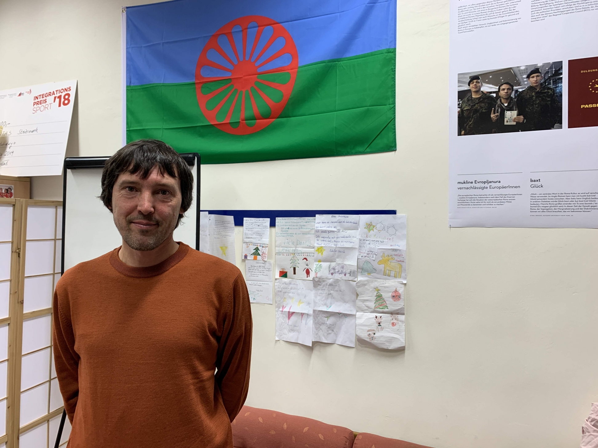 Michael Teichmann, Leiter des Projekt KAMBUKE bei der Caritas, steht vor einer Wand mit Flaggen und Kinderzeichnungen