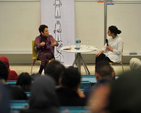 2 Frauen sitzen an einem Tisch und halten vor Studierenden einen Vortrag.