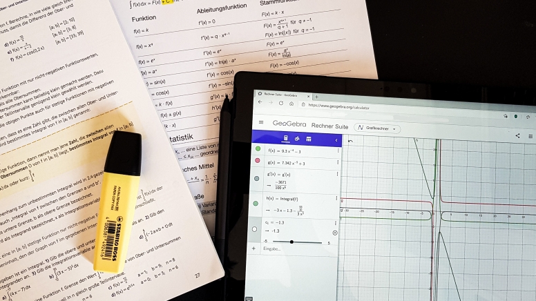 Arbeitsaufträge stapeln sich neben einem Mathematik-Programm am Tablet.