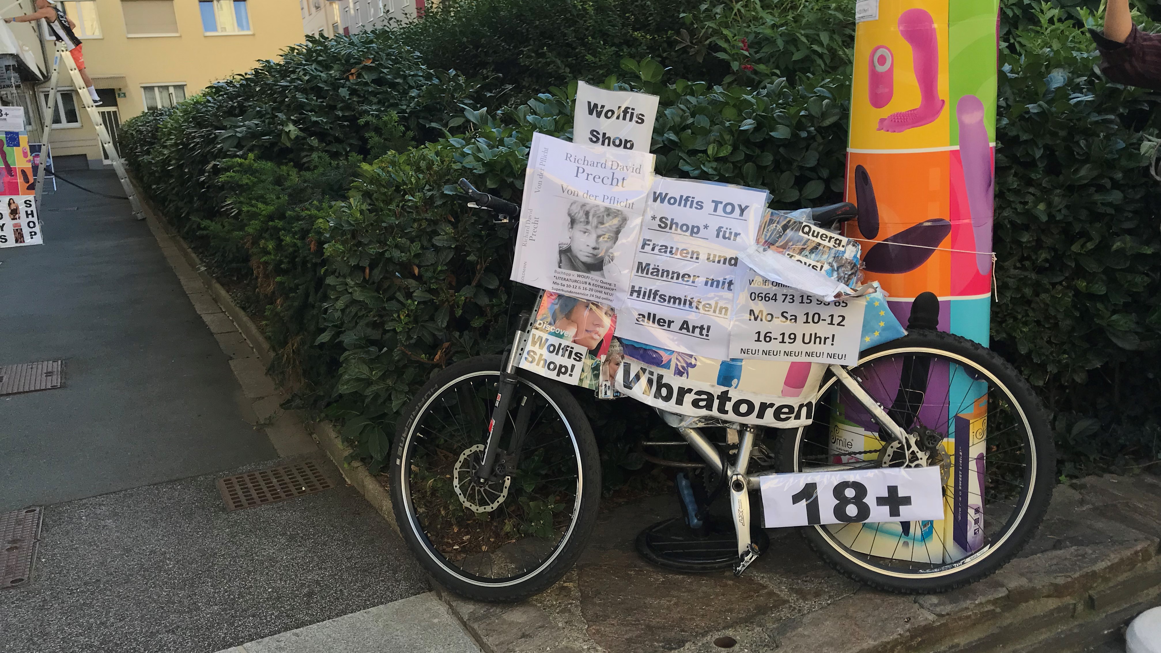 Ein Fahrrad, das an einer Hecke lehnt, auf dem sehr viele Zettel mit Werbung für den Erotikshop angebracht sind.