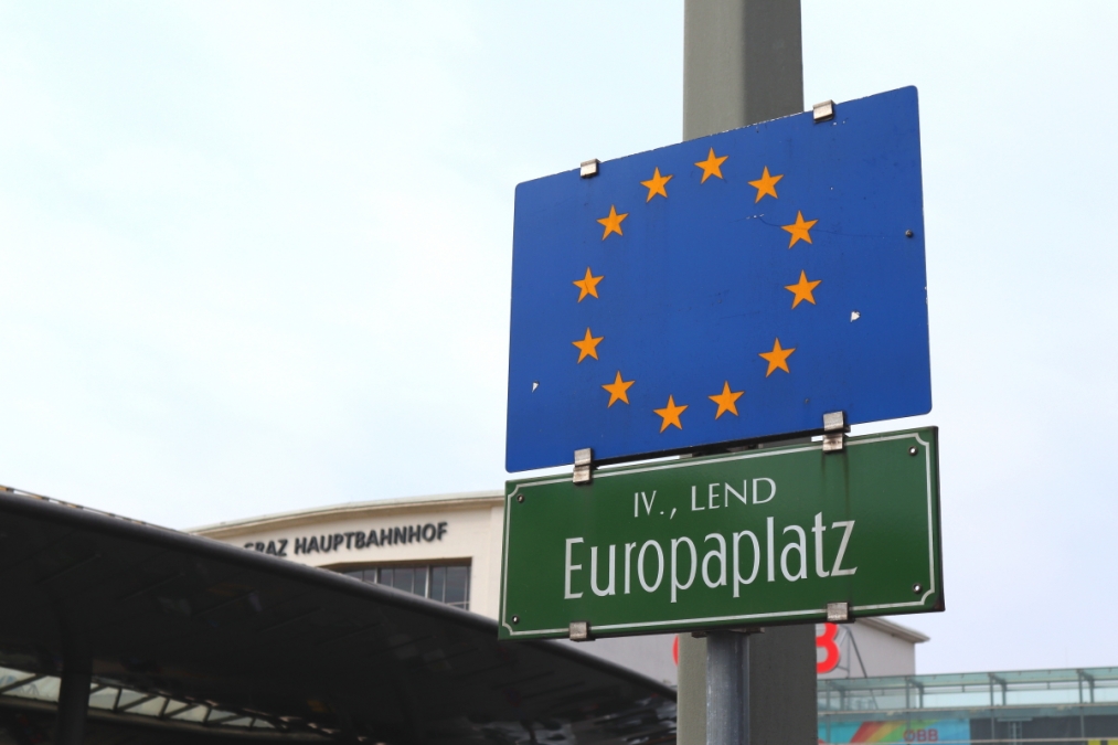 Dunkelgrünes Straßenschild am Europaplatz, darüber die EU-Fahne blau mit 12 gelben Sternen