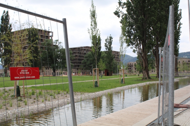 Die Wiese ist schon grün und das Wasserbecken eingelassen. Vor dem Park ist ein halboffener Bauzaun zu sehen.
