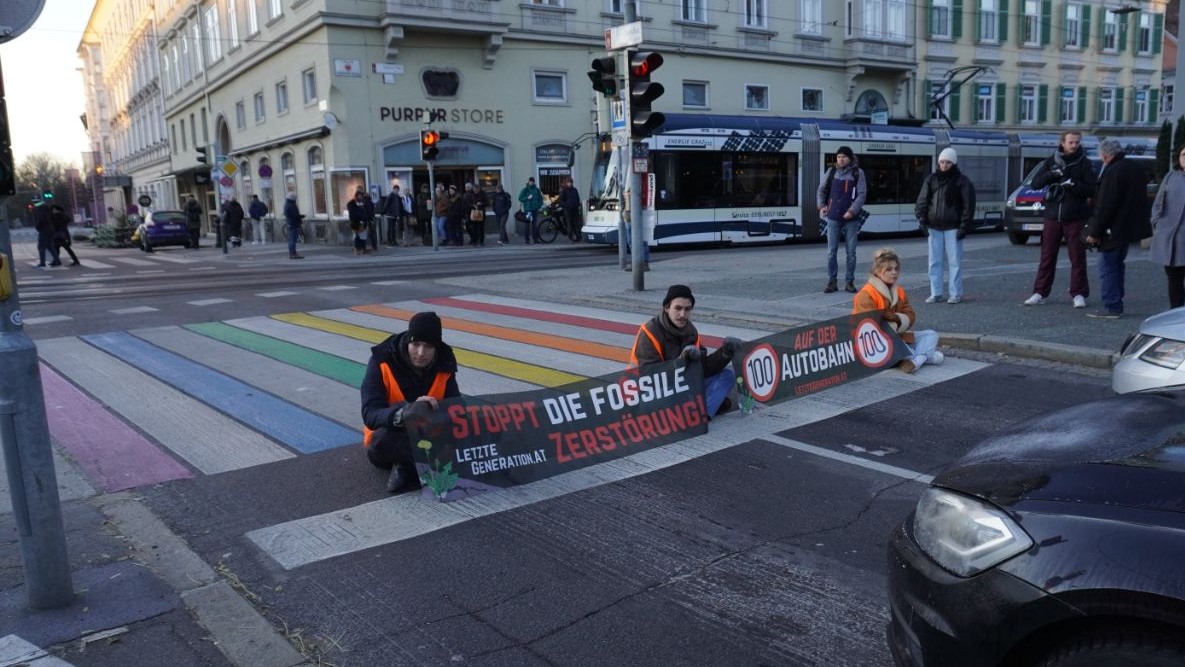 Drei Klimaaktivist:innen sitzen auf der Straße und halten Banner mit ihren Forderungen.