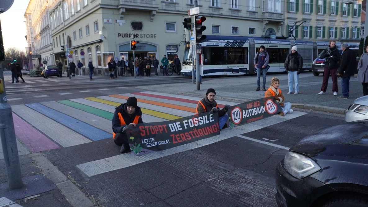 Drei Klimaaktivist:innen sitzen auf der Straße und halten Banner mit ihren Forderungen.
