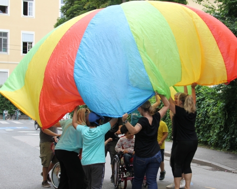 Unter einer Plane stehen Kinder des Mosaik Schulheims und Kinder des Schülerhorts Fröbelpark und spielen gemeinsam