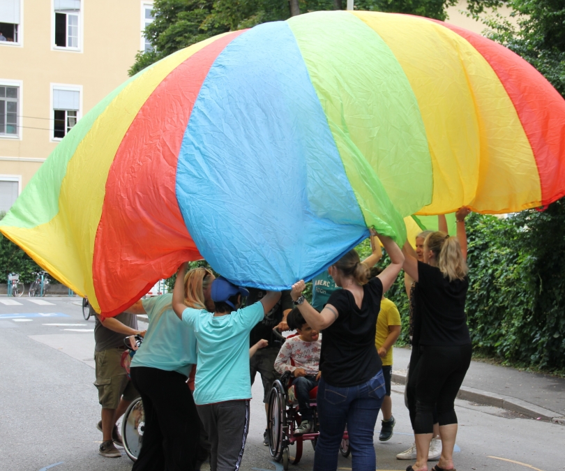 Unter einer Plane stehen Kinder des Mosaik Schulheims und Kinder des Schülerhorts Fröbelpark und spielen gemeinsam