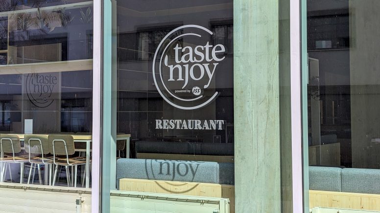 Das Logo von taste'njoy an einem der Fenster der Mensa.