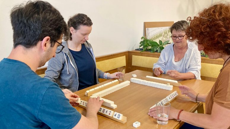 1 junger Mann mit Brille und 3 Frauen in unterschiedlichen Altersklassen sitzen um einen großen braunen Tisch. Auf diesem liegen weiße Steine mit Symbolen darauf. Sie spielen Mah.-Jongg.