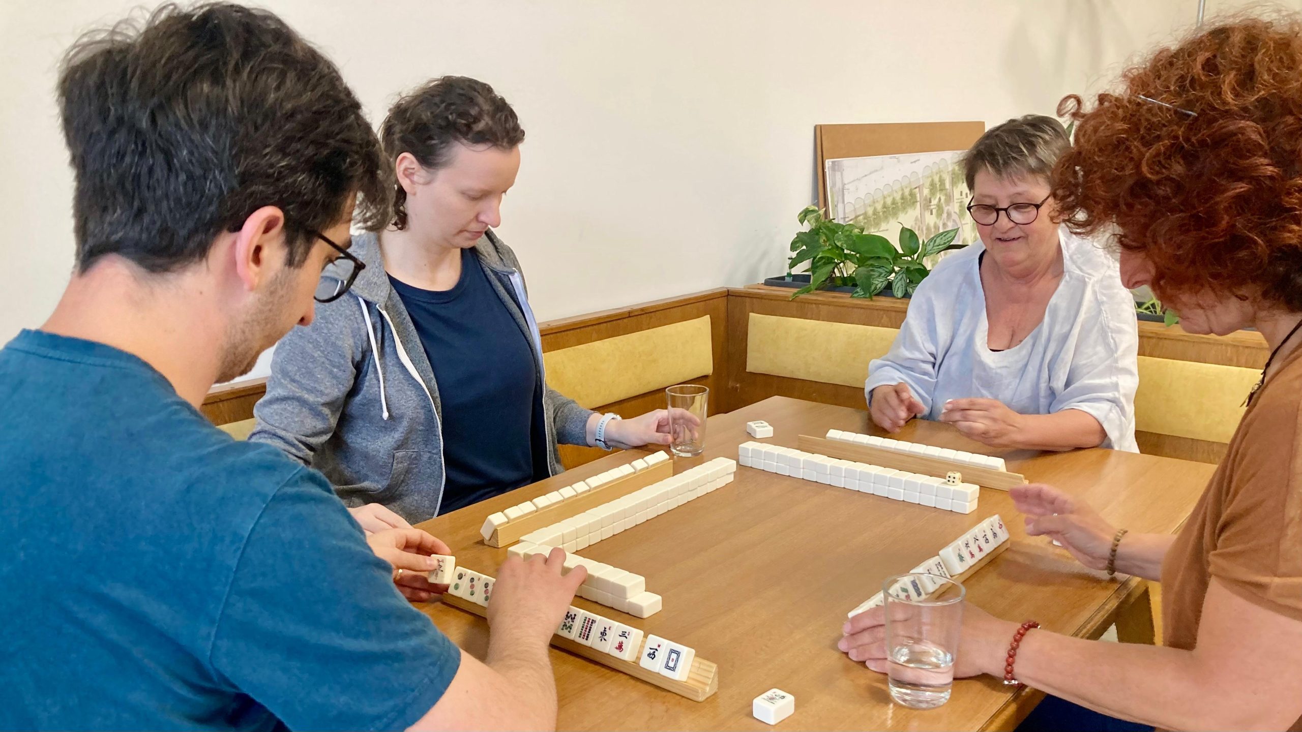 1 junger Mann mit Brille und 3 Frauen in unterschiedlichen Altersklassen sitzen um einen großen braunen Tisch. Auf diesem liegen weiße Steine mit Symbolen darauf. Sie spielen Mah.-Jongg.