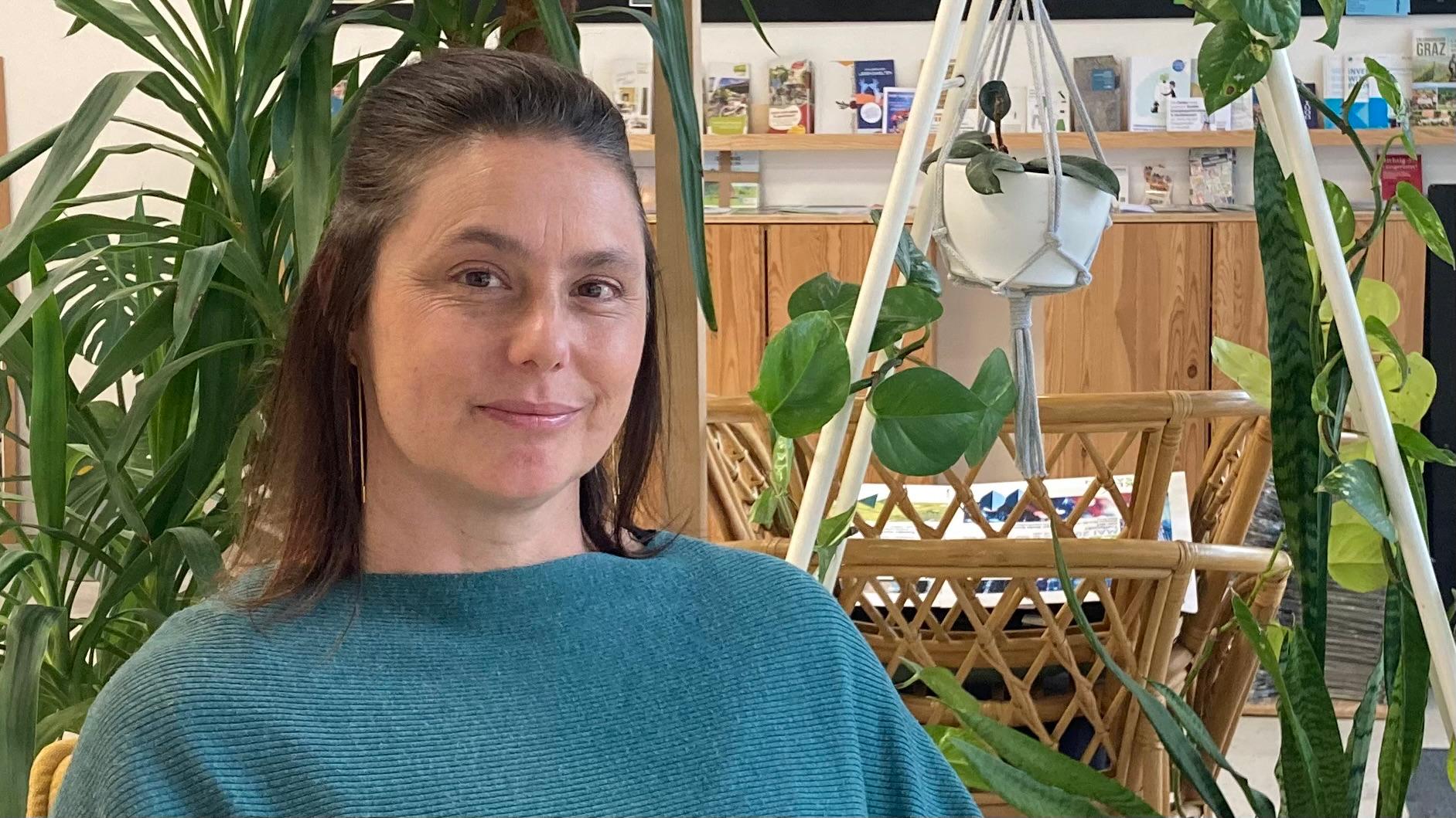 Eine Frau in einem blauen Pullover sitzt mittig und entspannt auf einem Polstersessel. Im Hintergrund sind grüne Pflanzen zu sehen.