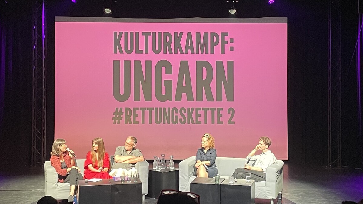 Zwei Moderator:innen und drei ungarische Kunstschaffende sitzen auf einer Couch und unterhalten sich vor einer rosa Leinwand mit der Aufschrift „KULTURKAMPF: UNGARN #RETTUNGSKETTE 2“.