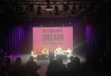 Ein Foto einer Podiumsdiskussion auf einer schwach beleuchteten Bühne. fünf Personen, zwei Moderator:innen und drei Kulturschaffende sitzen in hellen Sesseln auf der Bühne und sprechen. Hinter ihnen befindet sich eine große Leinwand, auf der in weißen Großbuchstaben auf rosa Hintergrund „KULTURKAMPF: UNGARN #RETTUNGSKETTE 2“ steht. Das Publikum ist im Vordergrund zu sehen, hauptsächlich von hinten.