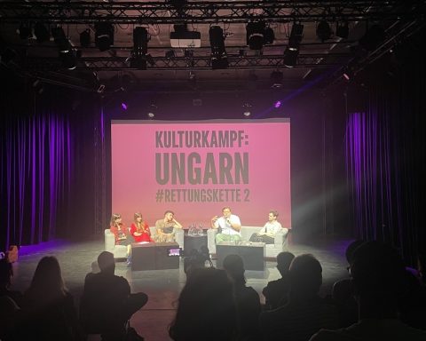 Ein Foto einer Podiumsdiskussion auf einer schwach beleuchteten Bühne. fünf Personen, zwei Moderator:innen und drei Kulturschaffende sitzen in hellen Sesseln auf der Bühne und sprechen. Hinter ihnen befindet sich eine große Leinwand, auf der in weißen Großbuchstaben auf rosa Hintergrund „KULTURKAMPF: UNGARN #RETTUNGSKETTE 2“ steht. Das Publikum ist im Vordergrund zu sehen, hauptsächlich von hinten.