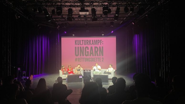 Ein Foto einer Podiumsdiskussion auf einer schwach beleuchteten Bühne. fünf Personen, zwei Moderator:innen und drei Kulturschaffende sitzen in hellen Sesseln auf der Bühne und sprechen. Hinter ihnen befindet sich eine große Leinwand, auf der in weißen Großbuchstaben auf rosa Hintergrund „KULTURKAMPF: UNGARN #RETTUNGSKETTE 2“ steht. Das Publikum ist im Vordergrund zu sehen, hauptsächlich von hinten.