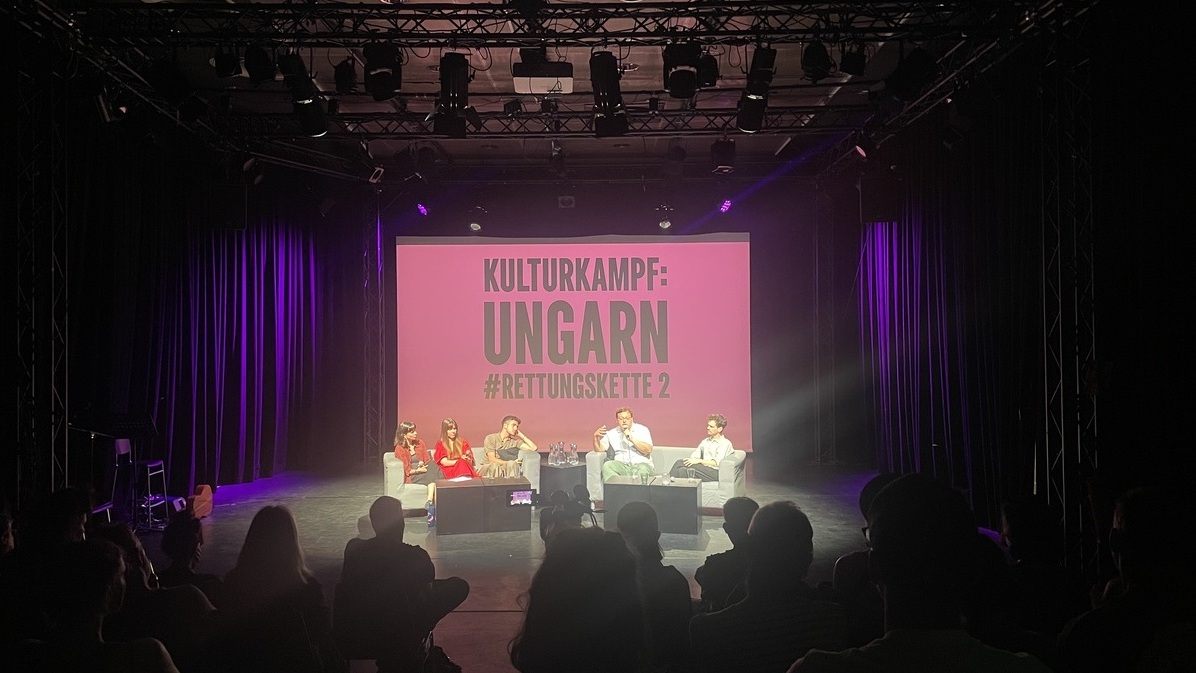 Ein Foto einer Podiumsdiskussion auf einer schwach beleuchteten Bühne. fünf Personen, zwei Moderator:innen und drei Kulturschaffende sitzen in hellen Sesseln auf der Bühne und sprechen. Hinter ihnen befindet sich eine große Leinwand, auf der in weißen Großbuchstaben auf rosa Hintergrund „KULTURKAMPF: UNGARN #RETTUNGSKETTE 2“ steht. Das Publikum ist im Vordergrund zu sehen, hauptsächlich von hinten.
