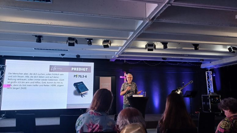 Ein Pastor steht neben einem großen Bildschirm, auf den Bibelverse zu sehen sind.