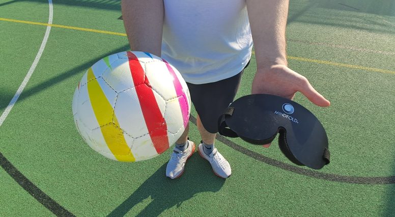 Eine Person steht auf einem grünen Outdoor-Sportplatz und hält einen bunten Blindenfußball in der linken Hand sowie eine schwarze Augenmaske mit der Aufschrift „Mindfold“ in der rechten. Der Ball ist weiß mit gelben, roten und pinken Streifen. Die Beine und Schuhe der Person sind sichtbar, der Oberkörper ist angeschnitten.