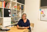 Gisela Kurath sitzt an ihrem Schreibtisch mit einem Bücherregal im Hintergrund.