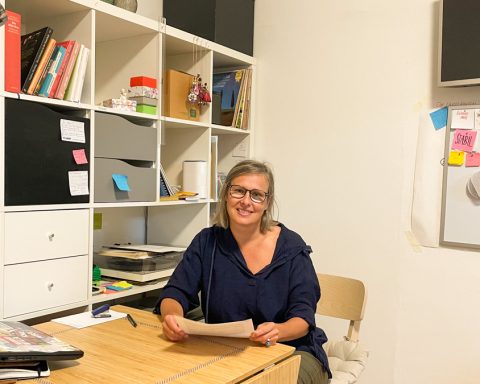 Gisela Kurath sitzt an ihrem Schreibtisch mit einem Bücherregal im Hintergrund.