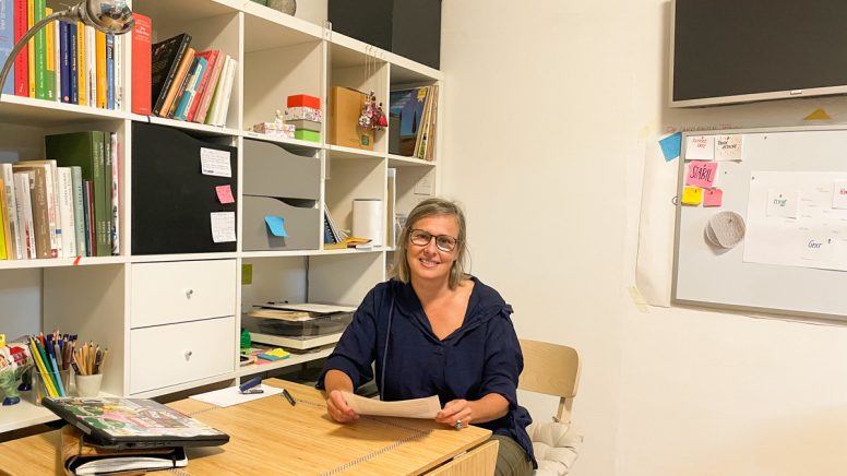 Gisela Kurath sitzt an ihrem Schreibtisch mit einem Bücherregal im Hintergrund.