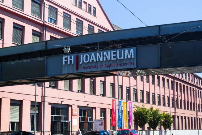 Außenansicht des Hauptgebäudes der FH JOANNEUM in Graz. Ein markanter Gehweg mit der Aufschrift ‚FH JOANNEUM – University of Applied Sciences‘ führt über die Straße. Das rosa-rote Gebäude im Hintergrund trägt mehrere bunte Fahnen mit dem Logo der Hochschule. Autos und Menschen befinden sich im unteren Bildbereich.