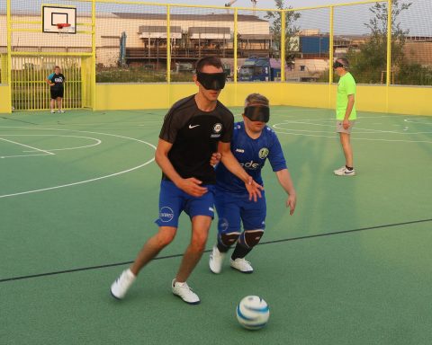 Zwei Fußballspieler mit Augenbinden kämpfen um einen Ball. Sie stehen auf einem Sportplatz mit grünem Belag und gelben Banden. Im Hintergrund stehen noch andere Spieler.