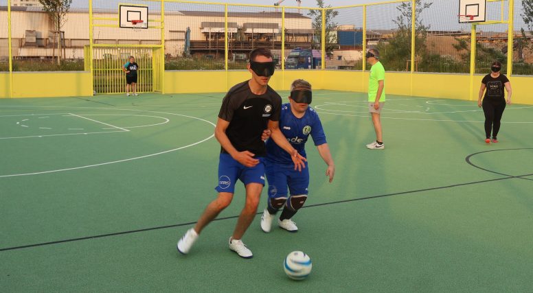 Zwei Fußballspieler mit Augenbinden kämpfen um einen Ball. Sie stehen auf einem Sportplatz mit grünem Belag und gelben Banden. Im Hintergrund stehen noch andere Spieler.
