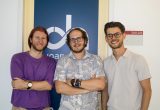 Drei junge Männer stehen nebeneinander und lächeln in die Kamera. Sie befinden sich vor einem dunkelblauen Schild mit dem Logo der FH JOANNEUM. Der Mann links trägt ein lila T-Shirt, der mittlere ein helles Hemd mit Blättermuster, der rechte ein helles Polohemd. Im Hintergrund ist ein Türschild mit der Raumnummer ‚EA11.222‘ zu sehen.