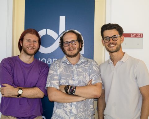 Drei junge Männer stehen nebeneinander und lächeln in die Kamera. Sie befinden sich vor einem dunkelblauen Schild mit dem Logo der FH JOANNEUM. Der Mann links trägt ein lila T-Shirt, der mittlere ein helles Hemd mit Blättermuster, der rechte ein helles Polohemd. Im Hintergrund ist ein Türschild mit der Raumnummer ‚EA11.222‘ zu sehen.
