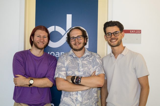 Drei junge Männer stehen nebeneinander und lächeln in die Kamera. Sie befinden sich vor einem dunkelblauen Schild mit dem Logo der FH JOANNEUM. Der Mann links trägt ein lila T-Shirt, der mittlere ein helles Hemd mit Blättermuster, der rechte ein helles Polohemd. Im Hintergrund ist ein Türschild mit der Raumnummer ‚EA11.222‘ zu sehen.