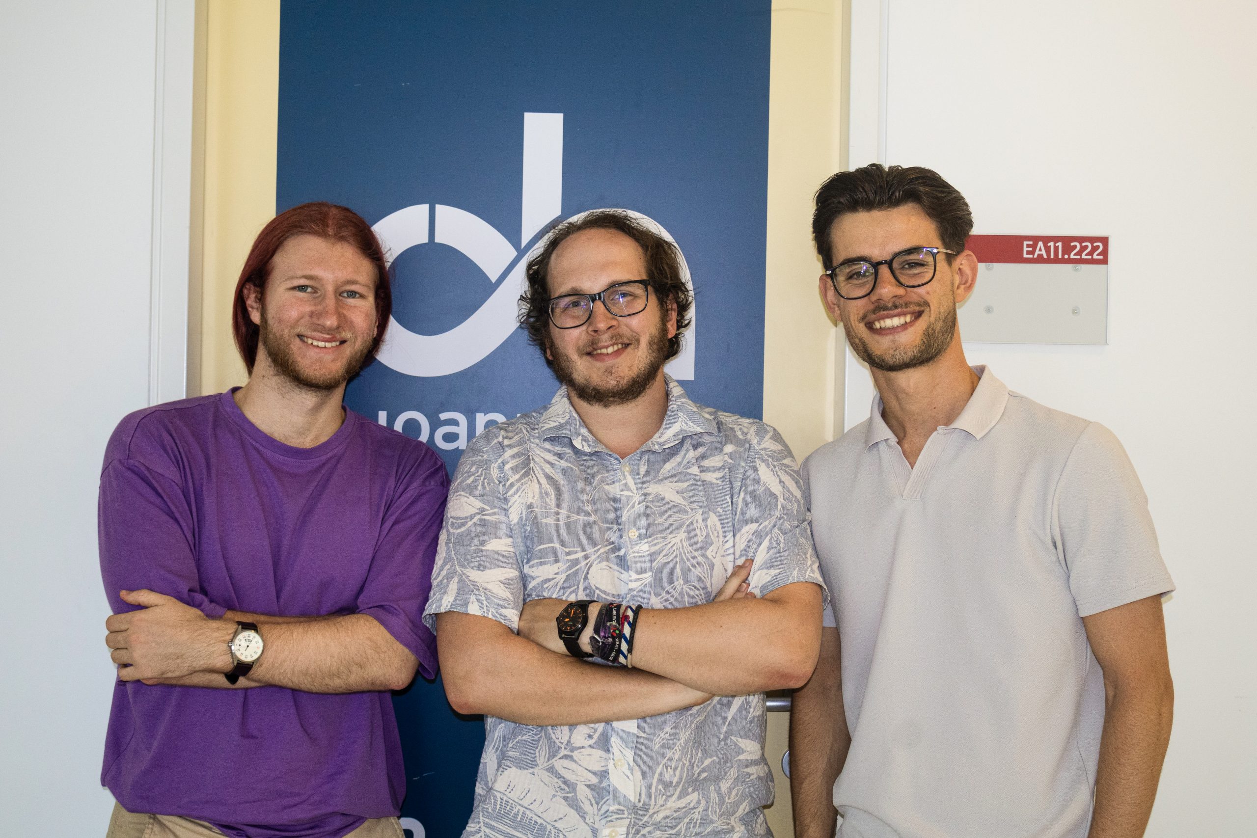 Drei junge Männer stehen nebeneinander und lächeln in die Kamera. Sie befinden sich vor einem dunkelblauen Schild mit dem Logo der FH JOANNEUM. Der Mann links trägt ein lila T-Shirt, der mittlere ein helles Hemd mit Blättermuster, der rechte ein helles Polohemd. Im Hintergrund ist ein Türschild mit der Raumnummer ‚EA11.222‘ zu sehen.