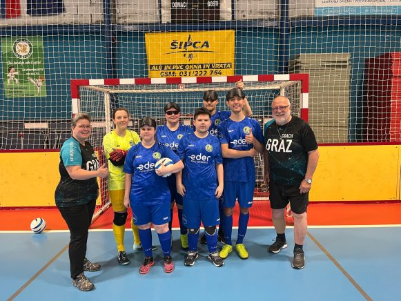 Ein gemischtes Blindenfußballteam posiert lächelnd in einer Sporthalle vor einem roten Tor mit Netz. Die Spieler:innen tragen blaue Trikots, einige haben Augenbinden. Zwei Trainer:innen stehen links und rechts außen, eine Person trägt ein gelbes Torwarttrikot. Ein Spieler hält den Ball. Alle wirken stolz und motiviert.