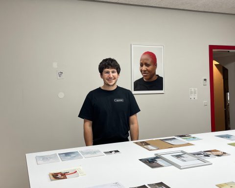 Elias Holzknecht steht in seiner Ausstellung vor einem Tisch mit Bildern. An der Wand hängt ein Porträt.