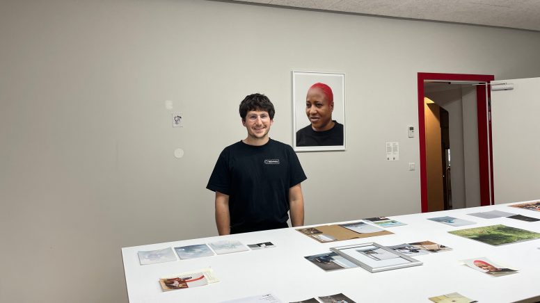 Elias Holzknecht steht in seiner Ausstellung vor einem Tisch mit Bildern. An der Wand hängt ein Porträt.