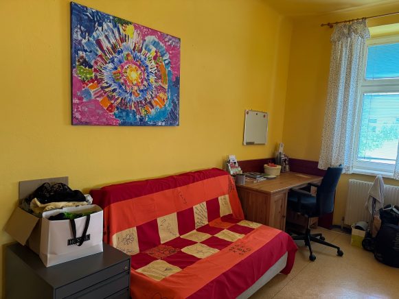 Rotes Sofa steht vor gelber Wand. Darüber hängt ein buntes Bild.