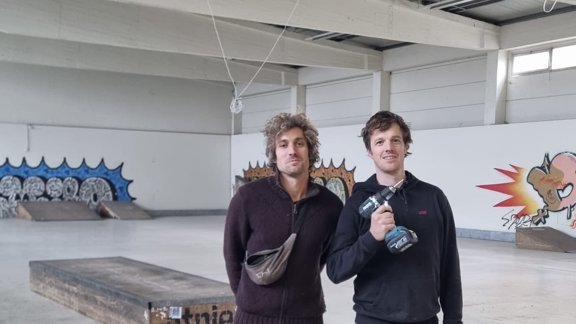 Zwei Männer stehen in der neuen Skatehalle vor buntem Graffiti. Der Mann links trägt eine weinrote Weste und eine braune Bauchtasche um seine Schulter. Der Rechte trägt einen dunkelblauen Kapuzenpullover und hält eine Bohrmaschine stolz hoch. Beide lächeln.