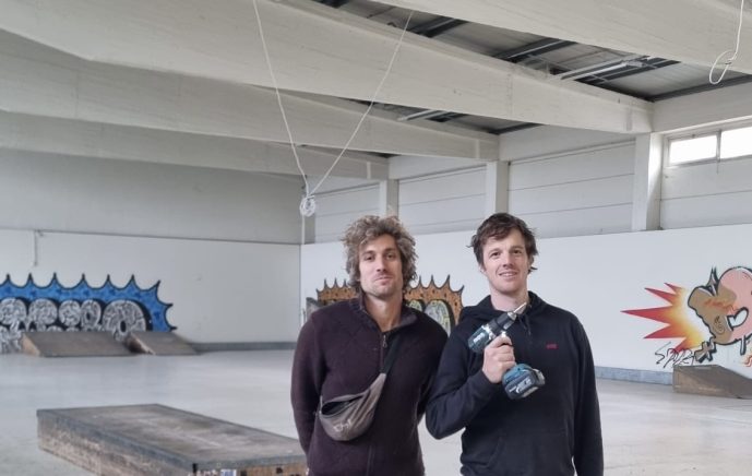 Zwei Männer stehen in der neuen Skatehalle vor buntem Graffiti. Der Mann links trägt eine weinrote Weste und eine braune Bauchtasche um seine Schulter. Der Rechte trägt einen dunkelblauen Kapuzenpullover und hält eine Bohrmaschine stolz hoch. Beide lächeln.