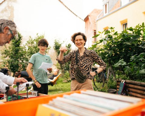 Zu sehen ist eine lachende Frau mit einem Jungen in einem Garten hinter einem Verkaufsstand.