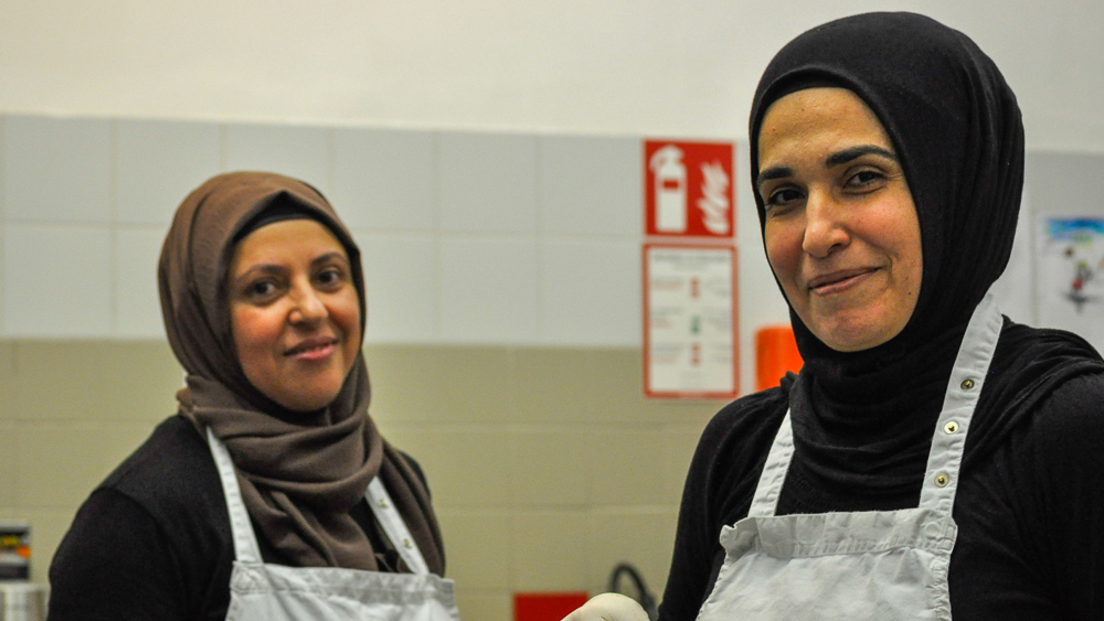 Die Küchengehilf:innen Meral Khan und Naile Kya stehen in der Küche und lächeln in die Kamera.