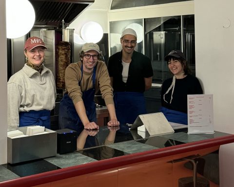 Auf dem Bild sind zwei Frauen und zwei Männer zu sehen, alle tragen eine Kopfbedeckung und lächeln in die Kamera. Im Hintergrund befindet sich ein Döner-Spieß.