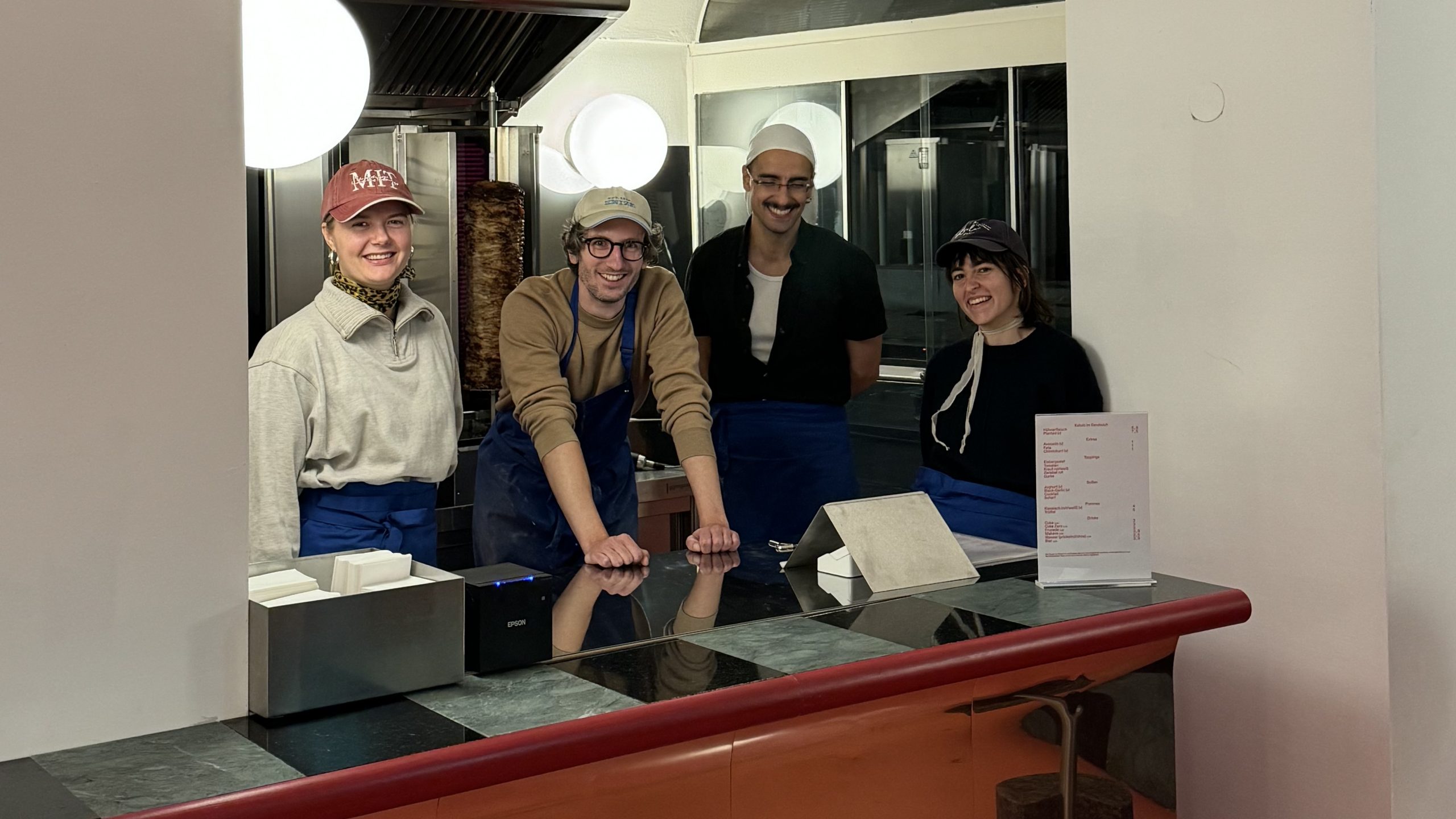 Auf dem Bild sind zwei Frauen und zwei Männer zu sehen, alle tragen eine Kopfbedeckung und lächeln in die Kamera. Im Hintergrund befindet sich ein Döner-Spieß.