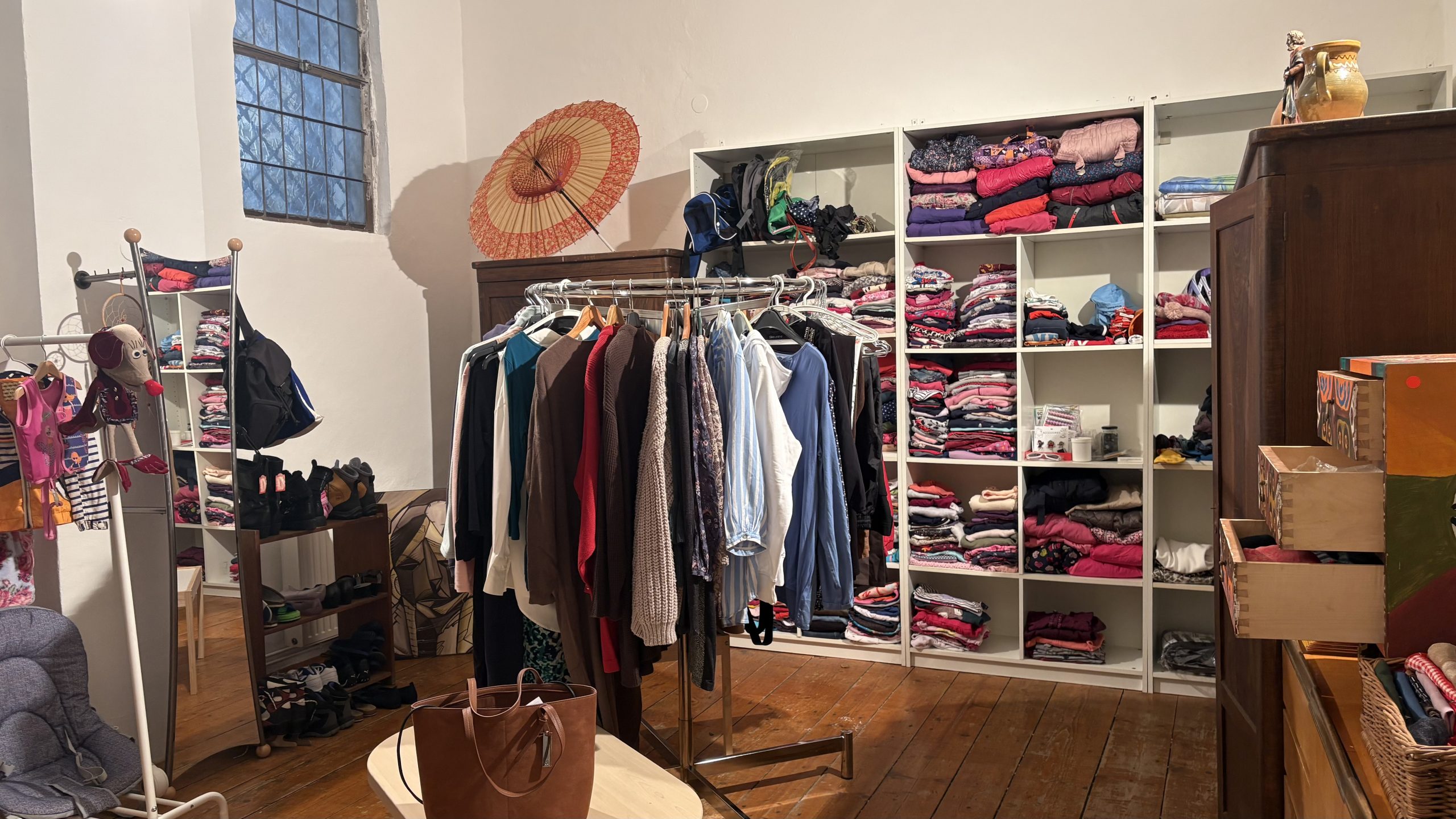 Zu sehen ist ein Regal, das mit Klamotten gefüllt ist sowie einige Kleiderständer.