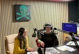 Zwei Frauen sitzen in einem Radio-Studio. In der Mitte der holzvertäfelten Wand hängt ein grünes Radio Helsinki Logo auf schwarzem Untergrund. Im Vordergrund sind Schaltpulte, Bildschirme und Mikrophone.