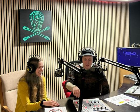 Zwei Frauen sitzen in einem Radio-Studio. In der Mitte der holzvertäfelten Wand hängt ein grünes Radio Helsinki Logo auf schwarzem Untergrund. Im Vordergrund sind Schaltpulte, Bildschirme und Mikrophone.