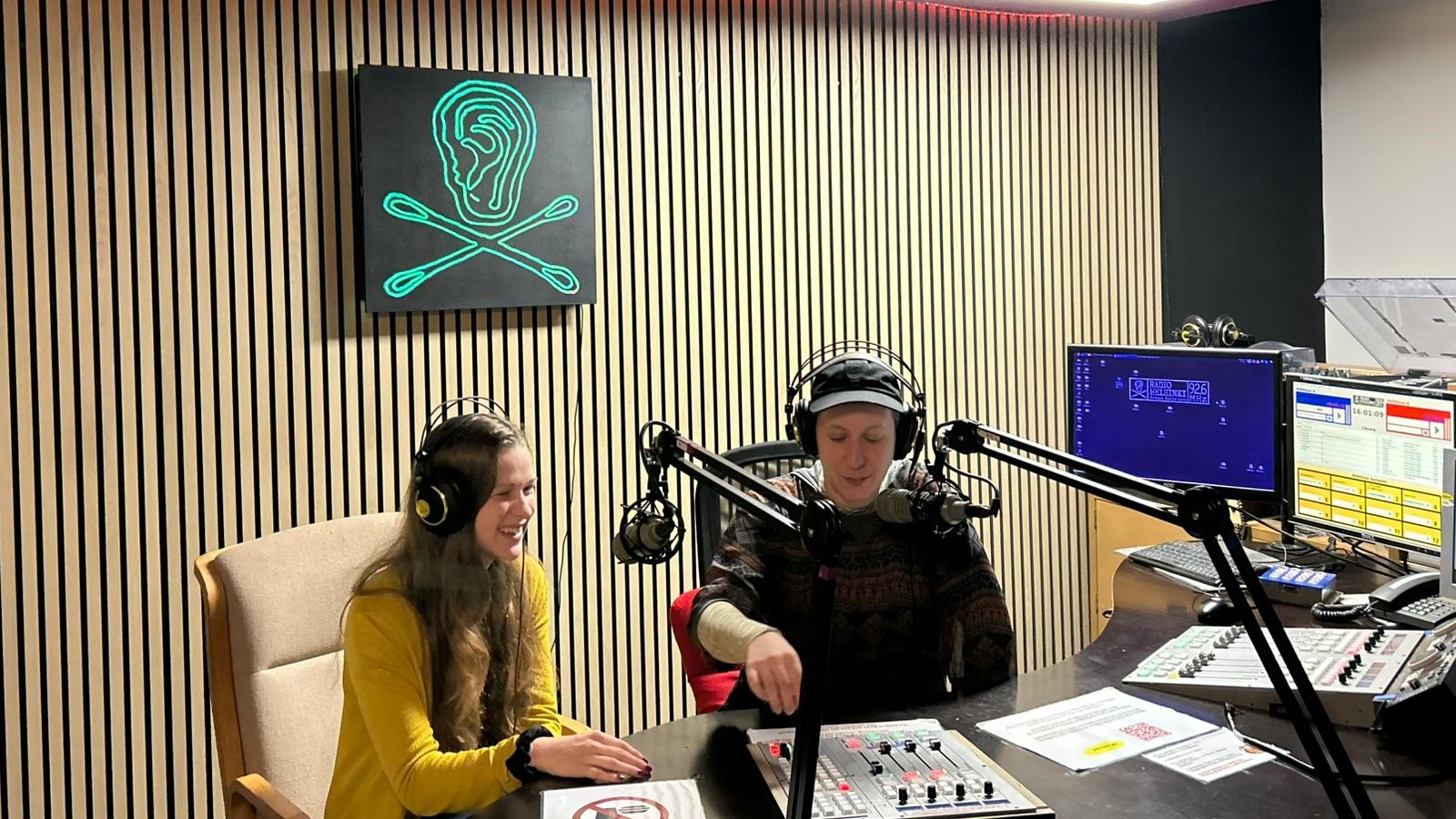 Zwei Frauen sitzen in einem Radio-Studio. In der Mitte der holzvertäfelten Wand hängt ein grünes Radio Helsinki Logo auf schwarzem Untergrund. Im Vordergrund sind Schaltpulte, Bildschirme und Mikrophone.