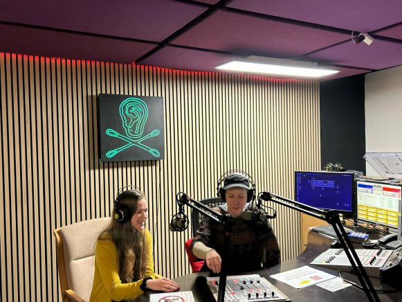 Zwei Frauen sitzen in einem Radio-Studio. In der Mitte der holzvertäfelten Wand hängt ein grünes Radio Helsinki Logo auf schwarzem Untergrund. Im Vordergrund sind Schaltpulte, Bildschirme und Mikrophone.