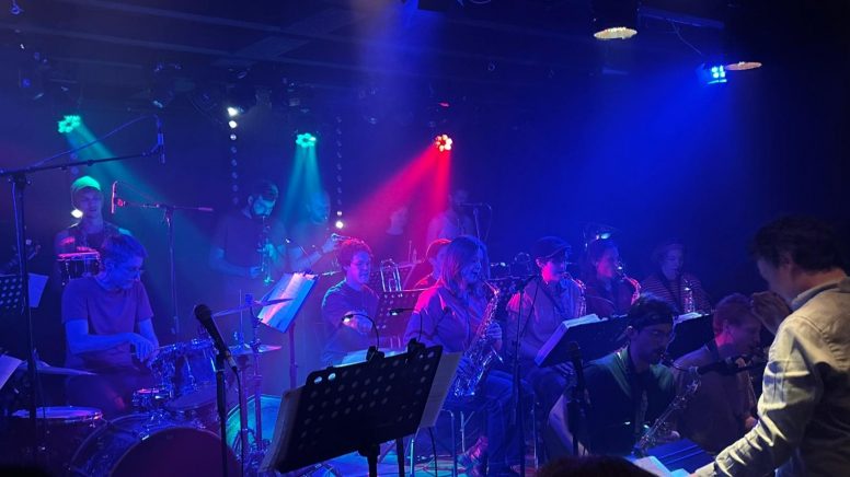 Viele Musiker stehen und sitzen auf einer Bühne. Alles ist in blaues Licht getaucht, ein roter Lichtstrahl durchbricht das Ganze.