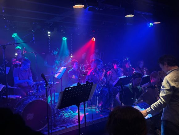 Viele Musiker stehen und sitzen auf einer Bühne. Alles ist in blaues Licht getaucht, ein roter Lichtstrahl durchbricht das Ganze.