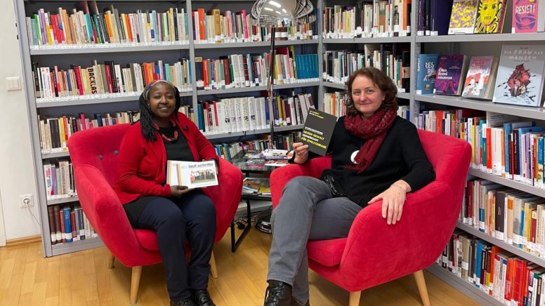 Auf dem Bild sind zwei Frauen vor einem großen Bücherregal mit feministischen Schriften. Die Frauen sind Petra Leschanz vom Verein Frauenservice und Edith Abawe vom Infocafe Palaver.